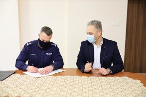 dwóch mężczyzn, jeden w stroju służbowym policjanta, drugi w marynarce, siedzą przy stole, podpisują dokument