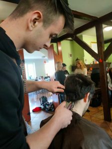 grupa ludzi, częśc stoi, część siedzi i korzysta z usług fryzjerskich