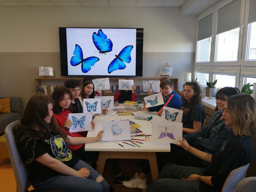 grupa młodych ludzi siedzi przy stole, w tle telewizor, ludzie trzymają kartki z niebieskimi motylami