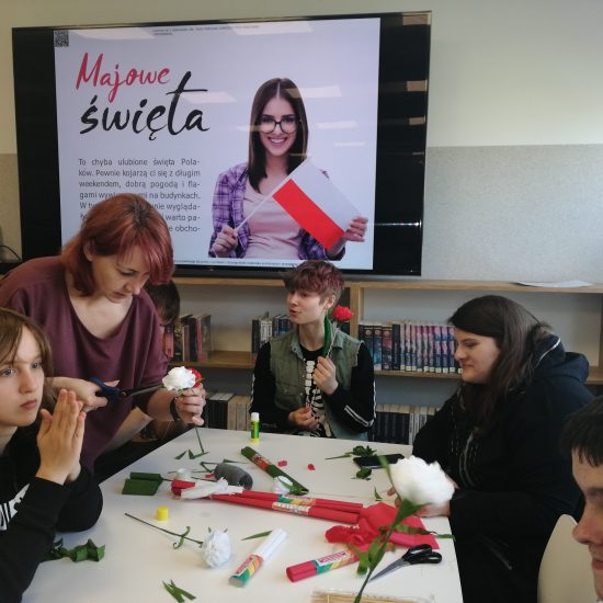 grupa ludzi siedzi przy stole, z bibuły robią kwiaty, goździki w kolorach czerwnonych i białych