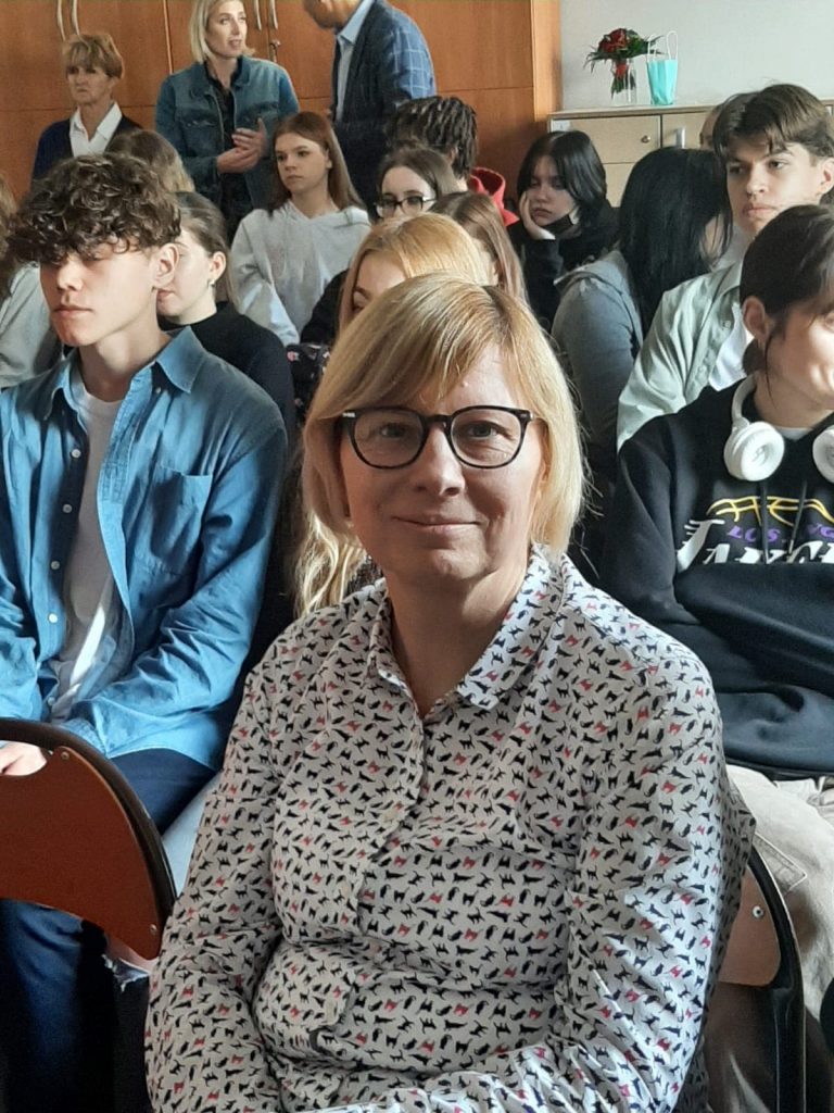 kobieta, blondynka w okularach, w tle grupa młodych osób, siedzą w sali lekcyjnej