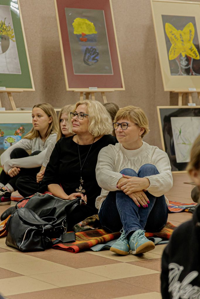 na pierwszym planie dwie kobiety w okularach, blondynki, krótkie włosy, za nimi dwie młode dziewczyny, siedzą na podłodze