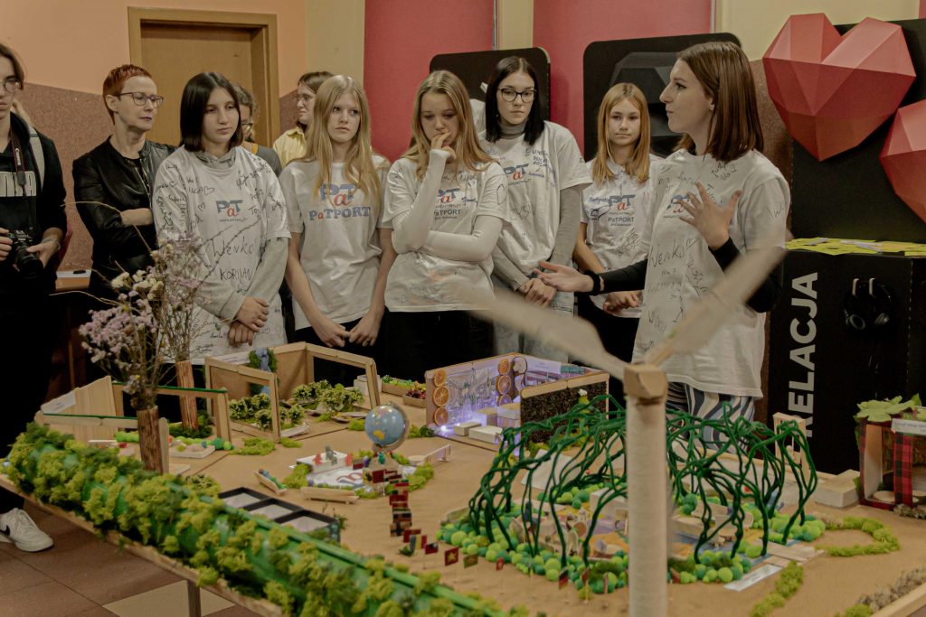 grupa młodych osób, dziewczyny w białych koszulkach, na pierwszym planie makieta, zielone elementy, mech, drewno