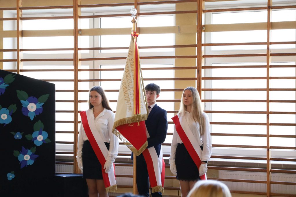trzy młode osoby, dwie dziewczyny po bokach, chłopak w środku trzyma sztandar, ubrani na galowo, szarfy z flagą Polski