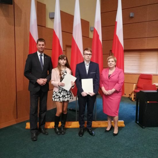 cztery osoby, z lewej mężczyzna w garniturze, dziewczyna w sukience z dyplomem, chłopak w garniturze, kobieta w różowej garsonce, w tle flagi Polski