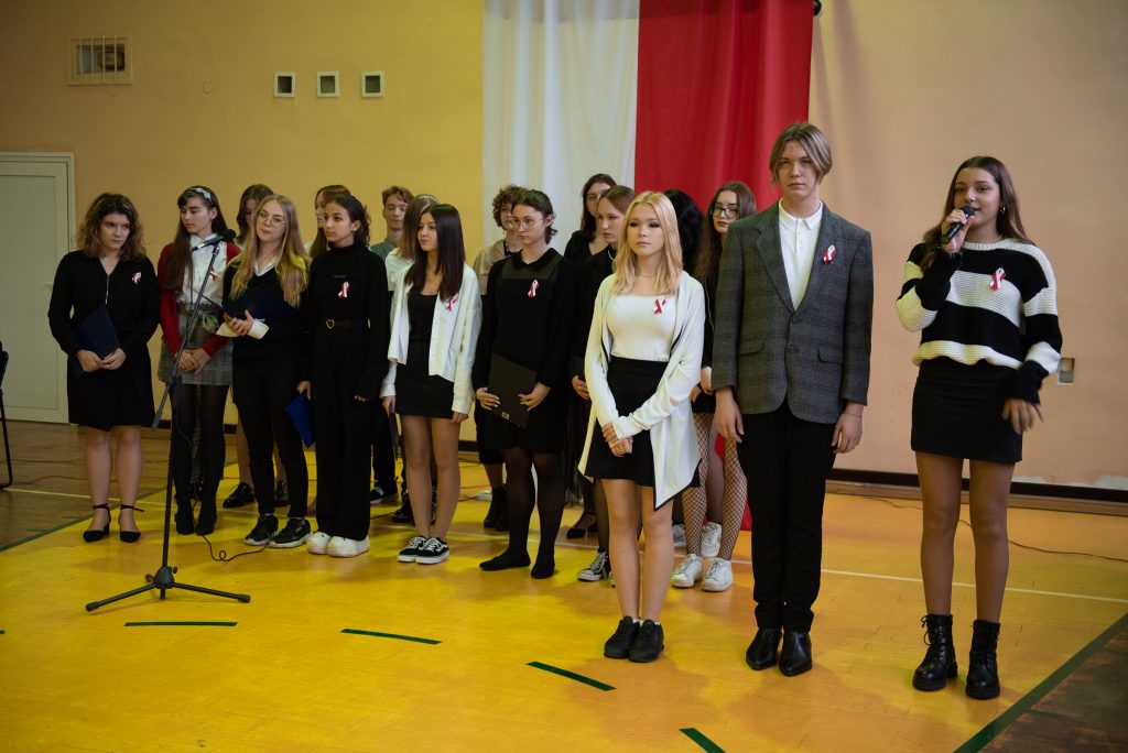 grupamłodych osób, chłopcy i dziewcyny, ubrani na galowo, mikrofony, w tle flaga Polski