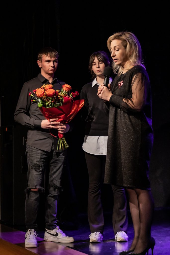 trzy osoby, chłopak trzyma bukiet kwiatów, kobieta, blondynka mówi do mikrofonu