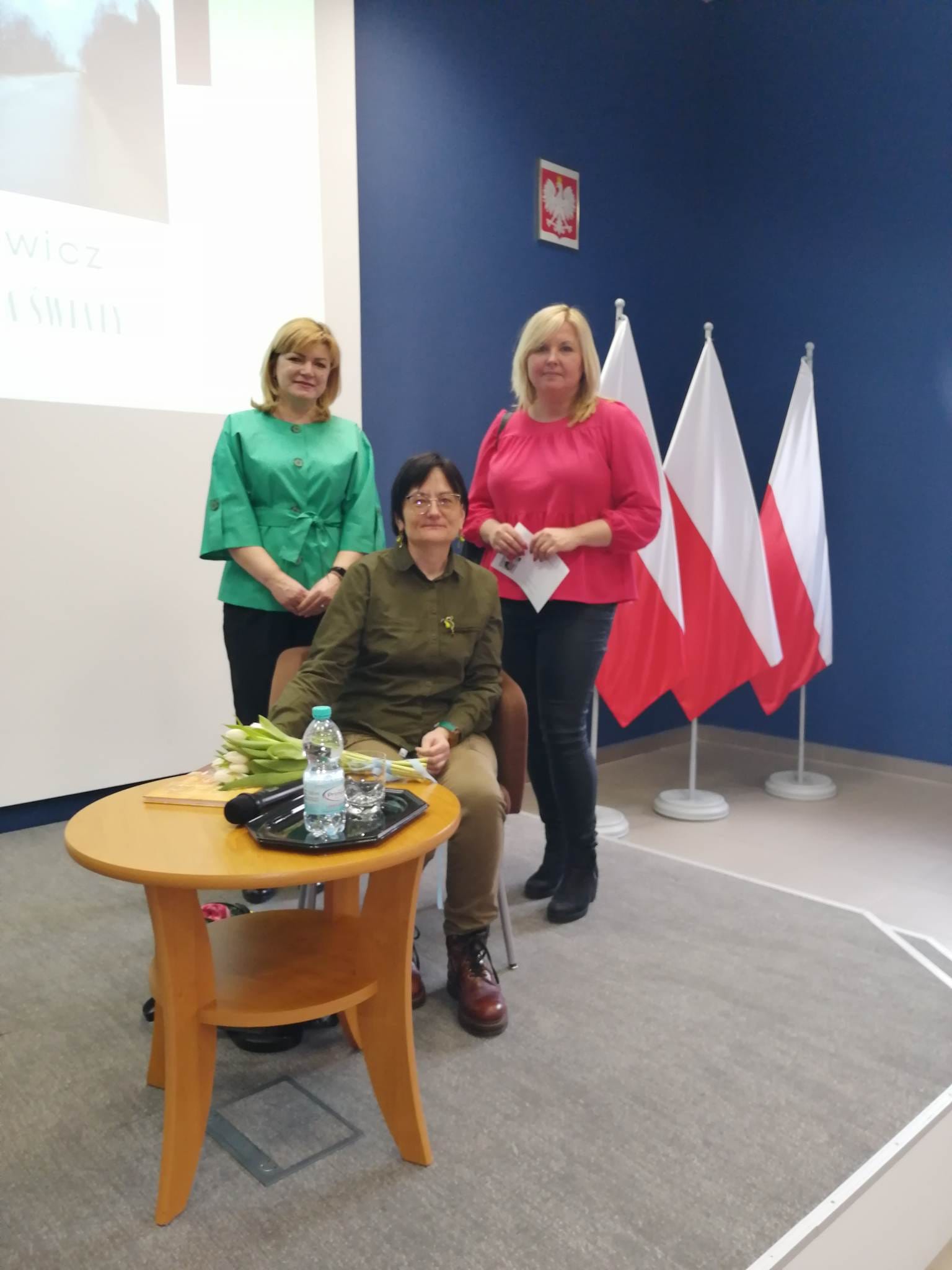 trzy kobiety, dwie stoją, jedna siedzi, w tle polskie flagi
