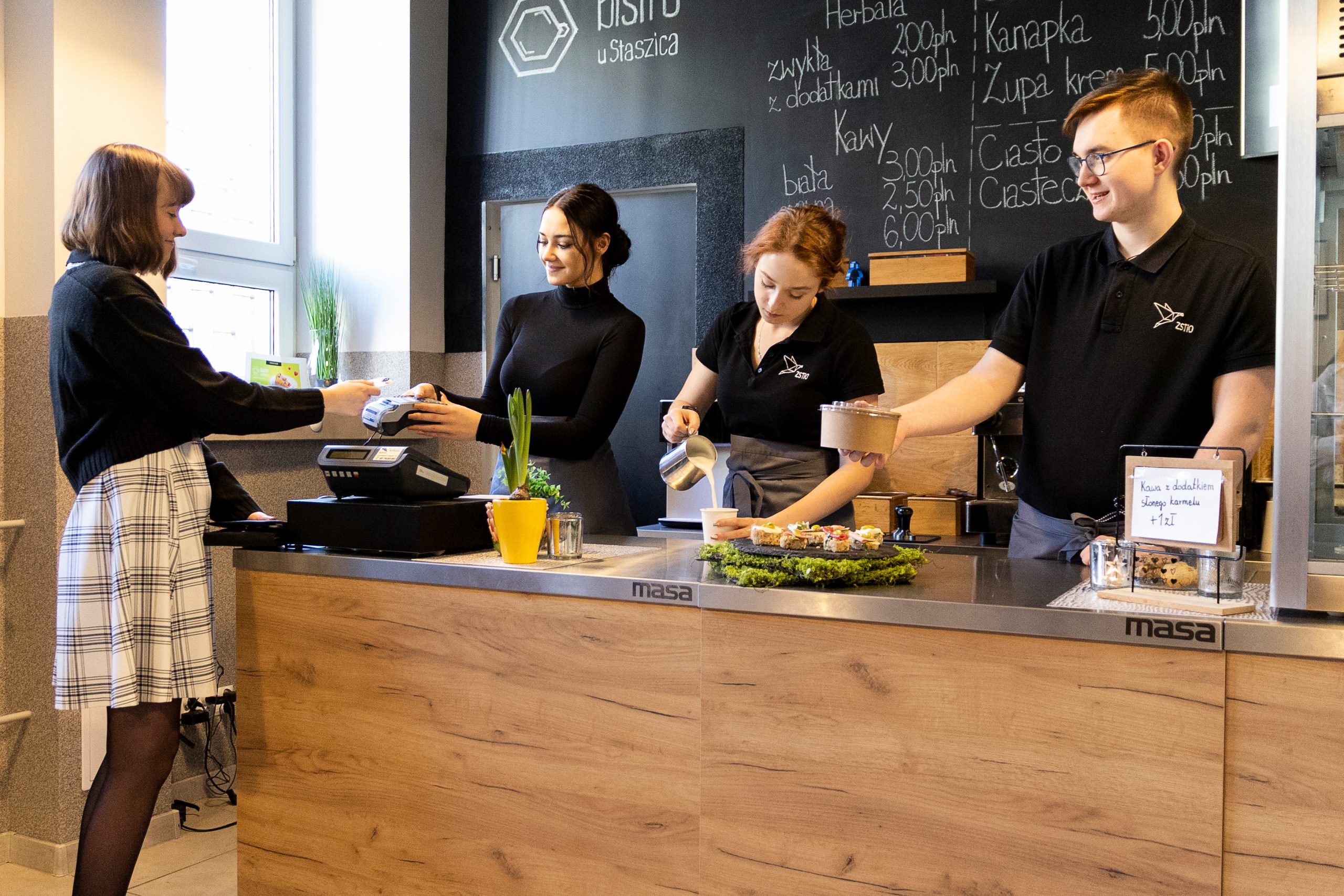 3 młdych kelnerów stoi za ladą , jeden, chłopak podaje sałatkę w pojemniku, dziewczyna leje mleko do kawy, dziewczyna z ciemnymi włosami podaje terminal płatniczy, dziewczyna stojąca tyłem opłaca kartą