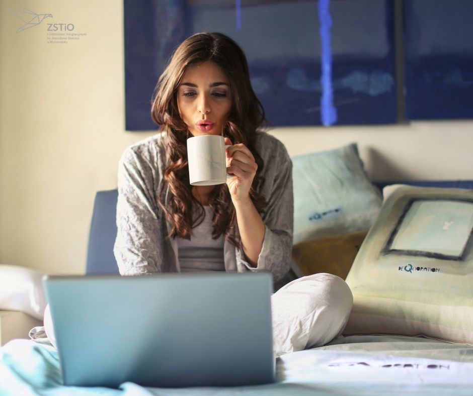 młoda dziewczyna siedzi na łóżku z laptopem, pije kawę, w tle poduszki.