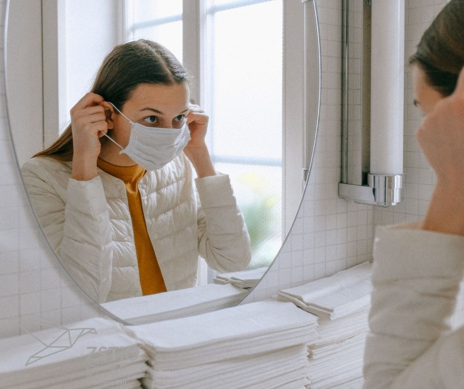 kobieta stoi w łazaience przed lusterkiem, zakłada maseczkę ochronną