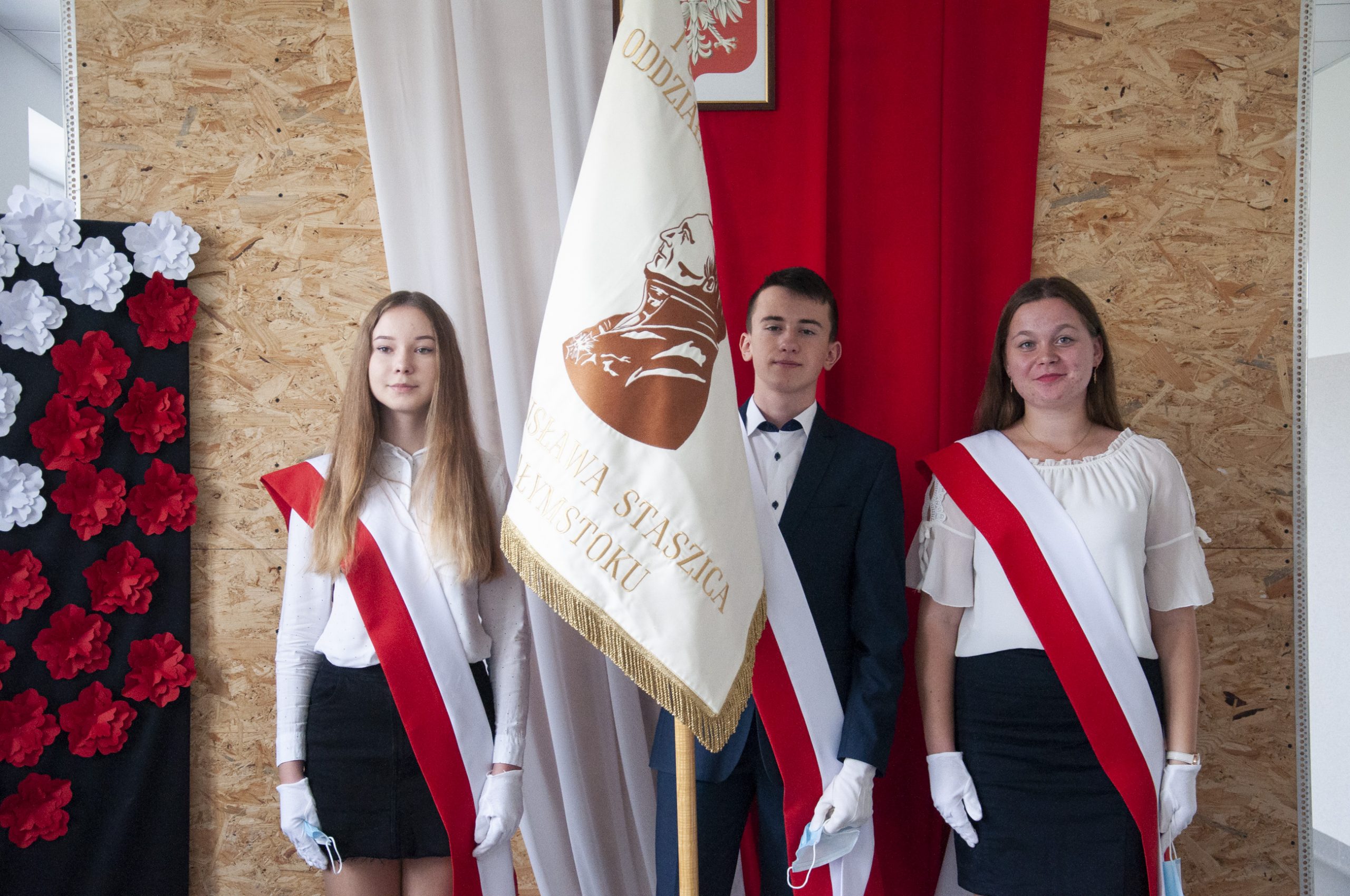 w tle flaga polska, trzy młode osoby w galowym stroju, chłopak trzyma sztandar szkoły