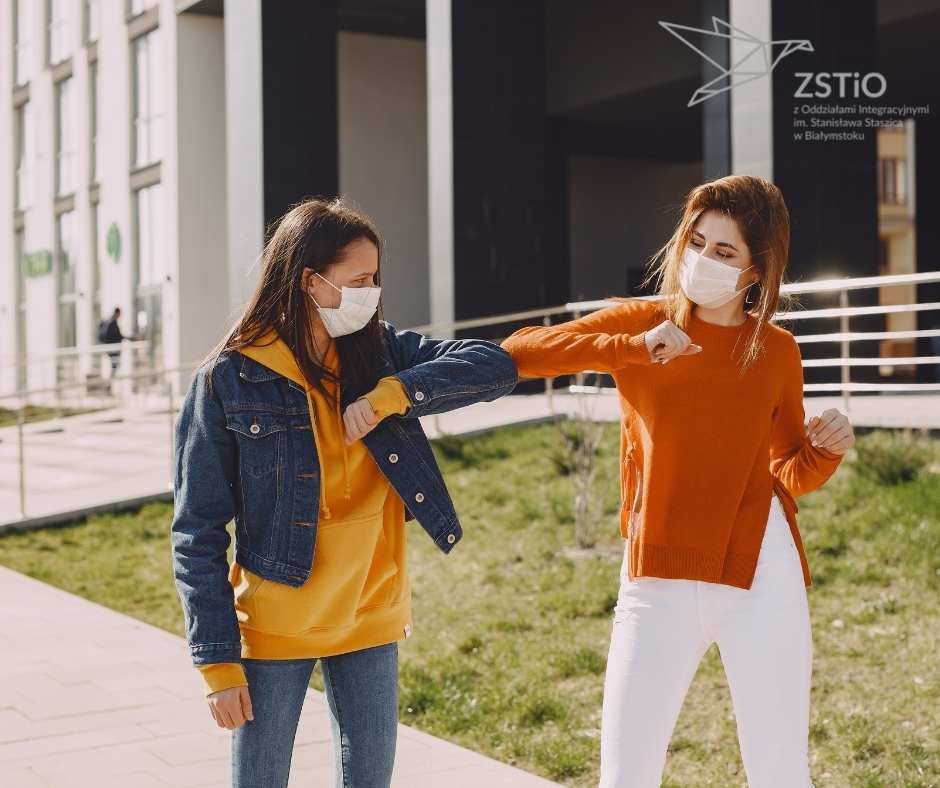 dwie młode kobiety w maseczkach, witają się łokciami