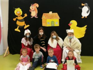 w tle zwierzęta, na pierwdszym planie siedzą dzieci, z tyłu cztery dziewczyny przebrane w historyczne stroje