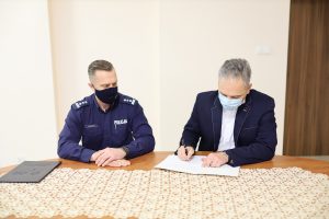 dwóch mężczyzn, jeden w stroju służbowym policjanta, drugi w marynarce, siedzą przy stole, podpisują dokument