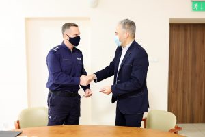 dwóch mężczyzn, jeden w stroju służbowym policjanta, drugi w marynarce, siedzą przy stole, uścisk dłoni