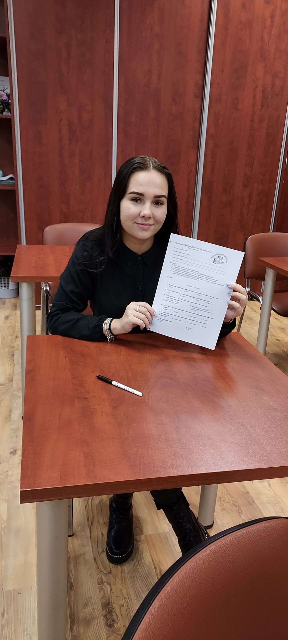 młoda dziewczyna ubrana na czarno siedzi w szkolnej ławce, w rękach trzyma kartkę z testem