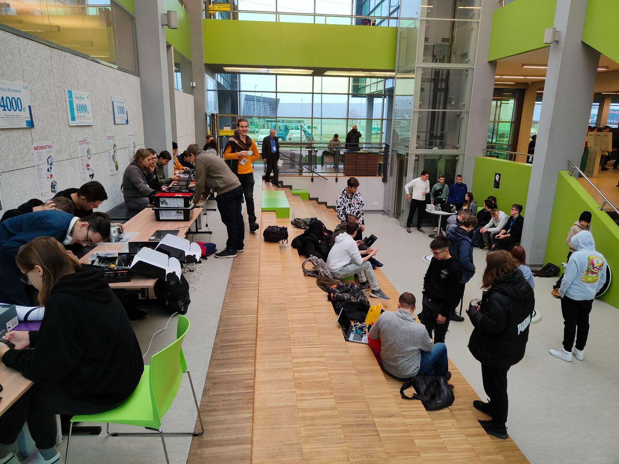 aula, drewniana podłoga, zielone lelementy, ludzie siedzą przy stołach i na schodach