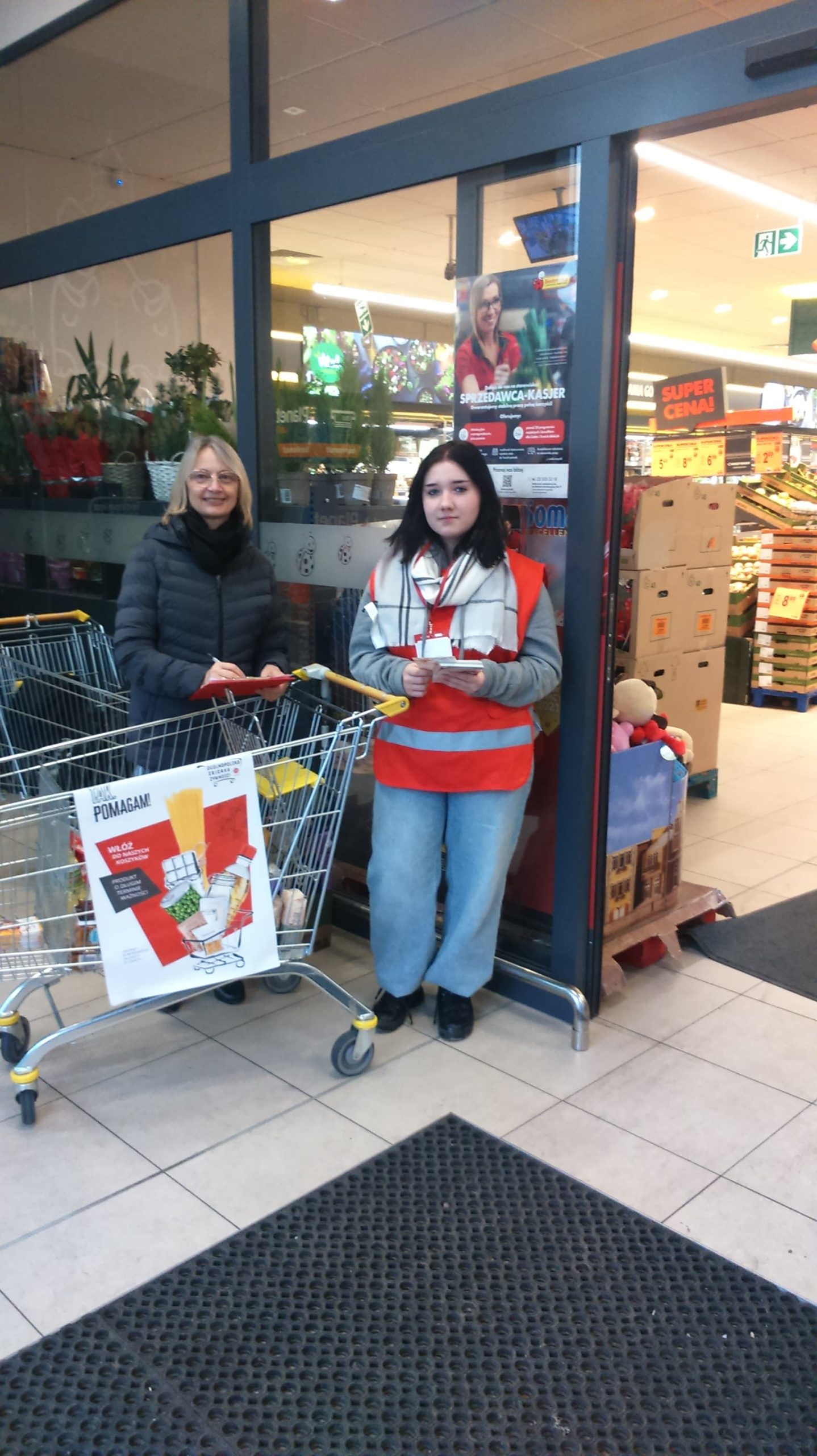 dwie kobiety stoją przed sklepem, jedna tyrzyma wózek sklepowy, wolontariuszki