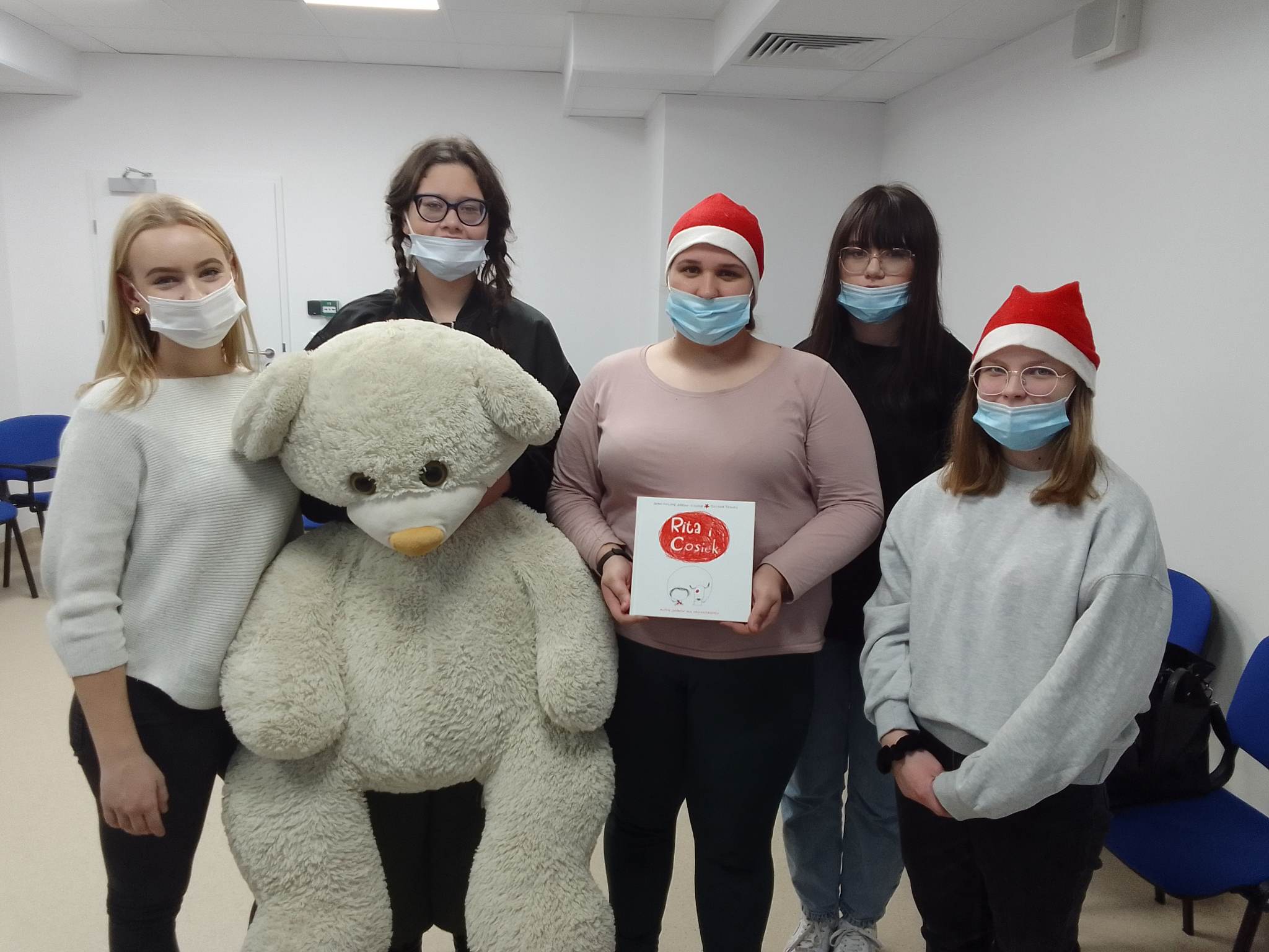 5 dziewczyn, dwie w czapkach mikołaja, jedna ytrzyma dużego pluszowego misia, wszystkie w maseczkach
