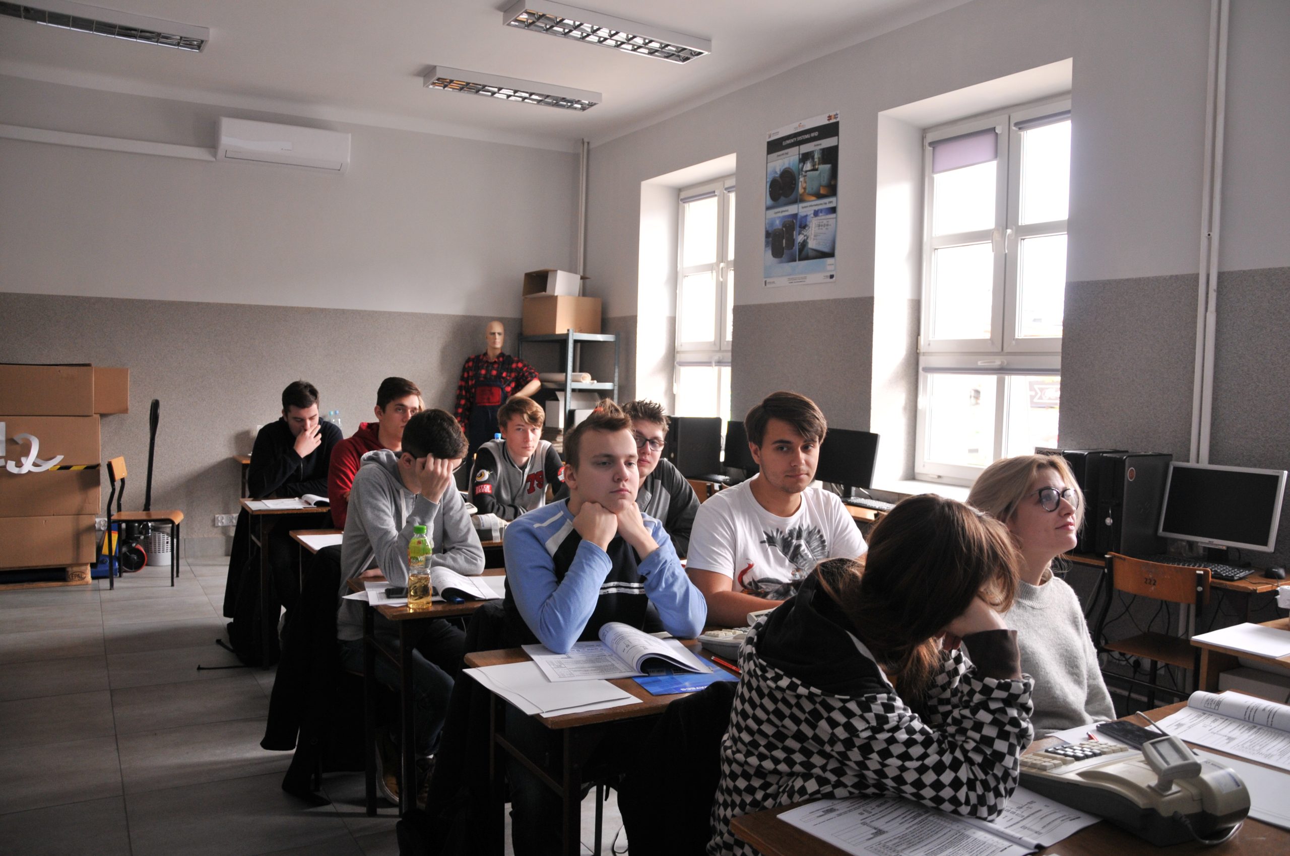 uczniowie siedzący w ławkach, w tle jasne okna