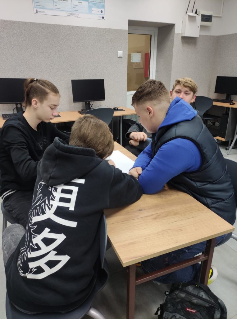 4 uczniów siedzi przy biurku, na stole piórniki, mazaki, kredki, kartki