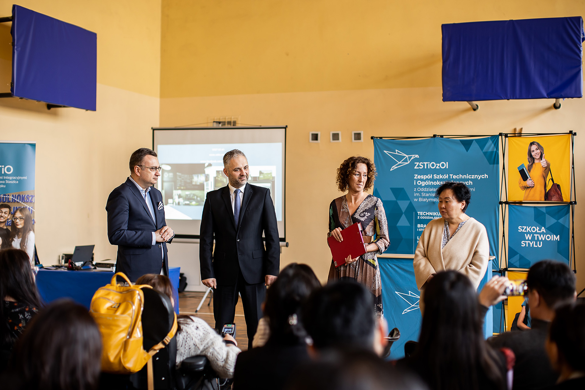 dwóch mężczyzn w garniturze, kobieta w sukience i kobieta w beżowym swetrze stoja na sali gimnastycznej