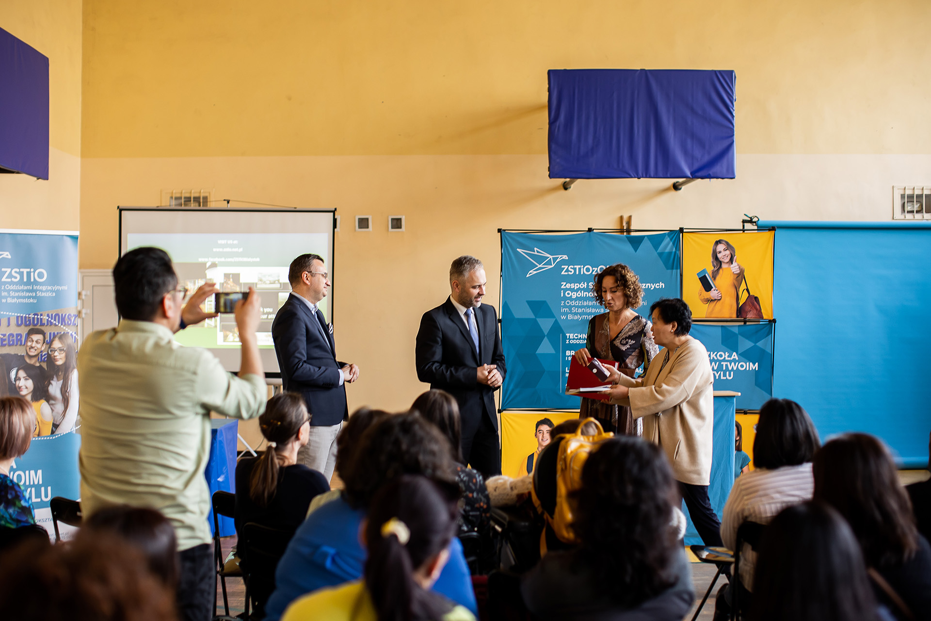 kobieta w bezowym swetrze wręcza ptrezent mężczyźnie w garniturze, obok mężczzyzna w garniturze
