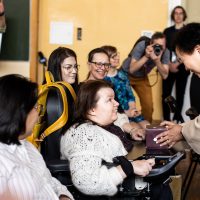 kobieta na wózku inwalidzkim dostaje prezent od kobiety w beżowym swetrze, w tle inni ludzie siedzą na krzesłach