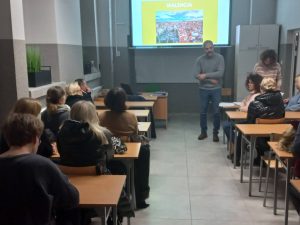 mężczyzna w swetrze, w sali ludzie siedzą w ławkach szkolnych