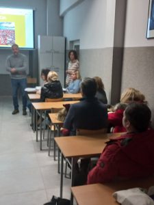 mężczyzna w swetrze, w sali ludzie siedzą w ławkach szkolnych