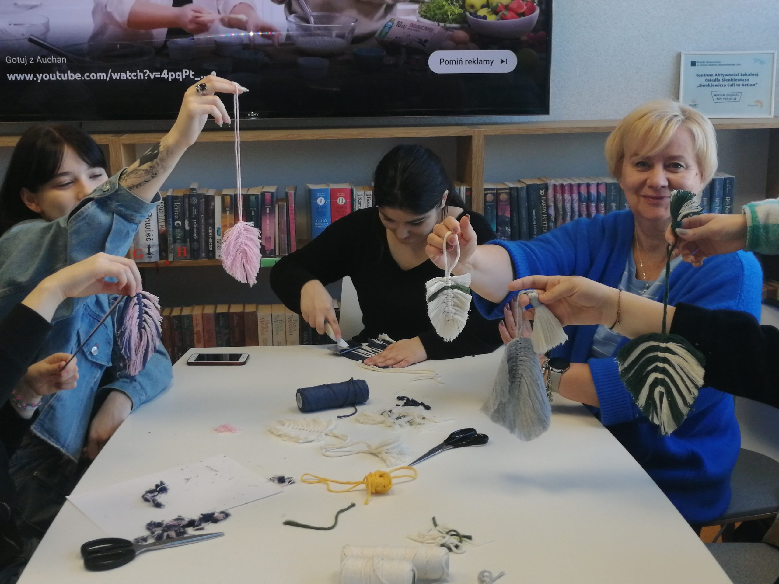 grupa ludzi w różnym wieku siedzi przy stole i robią makramy