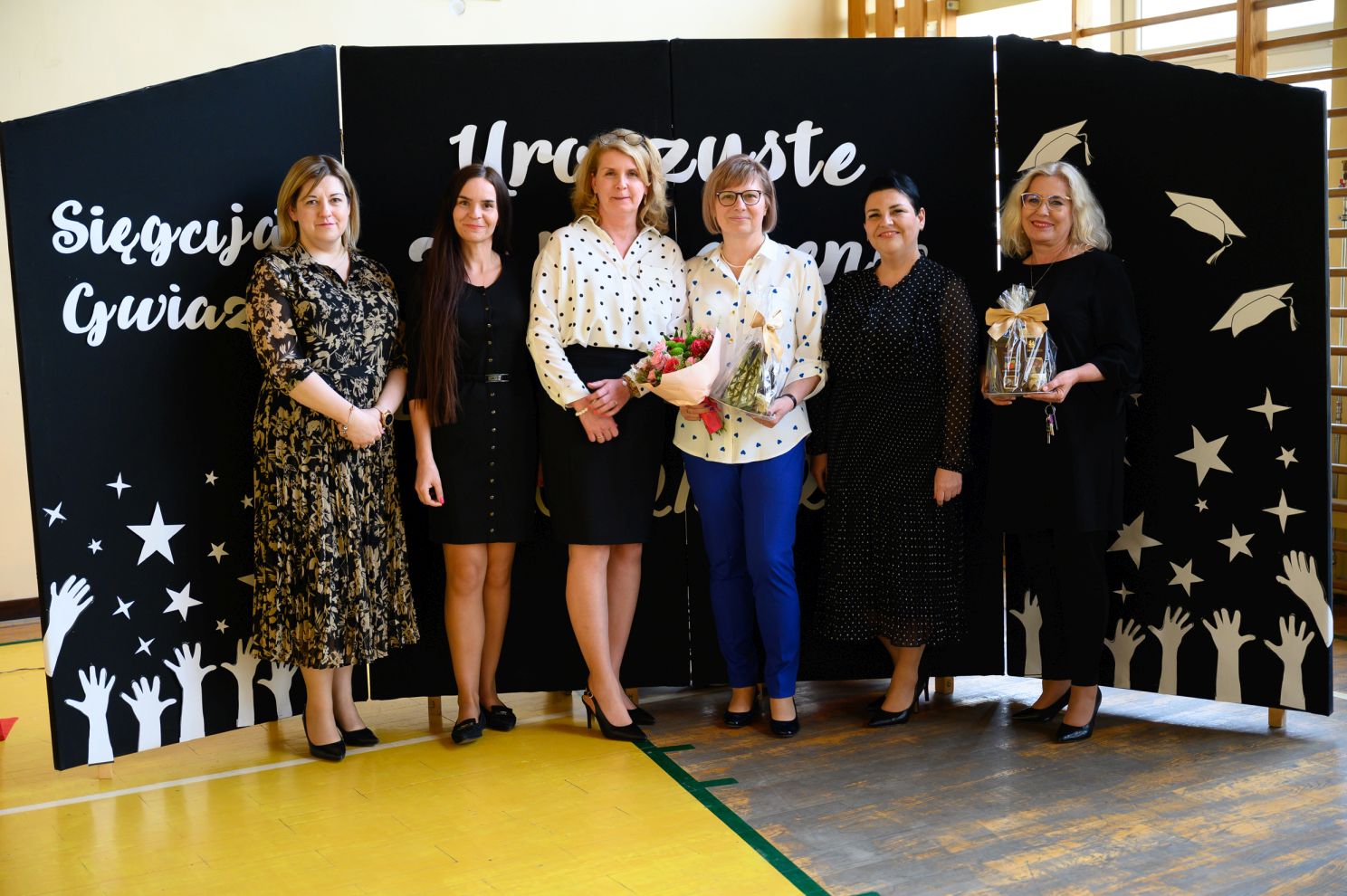 6 kobiet ubranych w sukienki pozuje do zdjecia, dwie blondynki trzymają kosze prezentowe i kwiaty