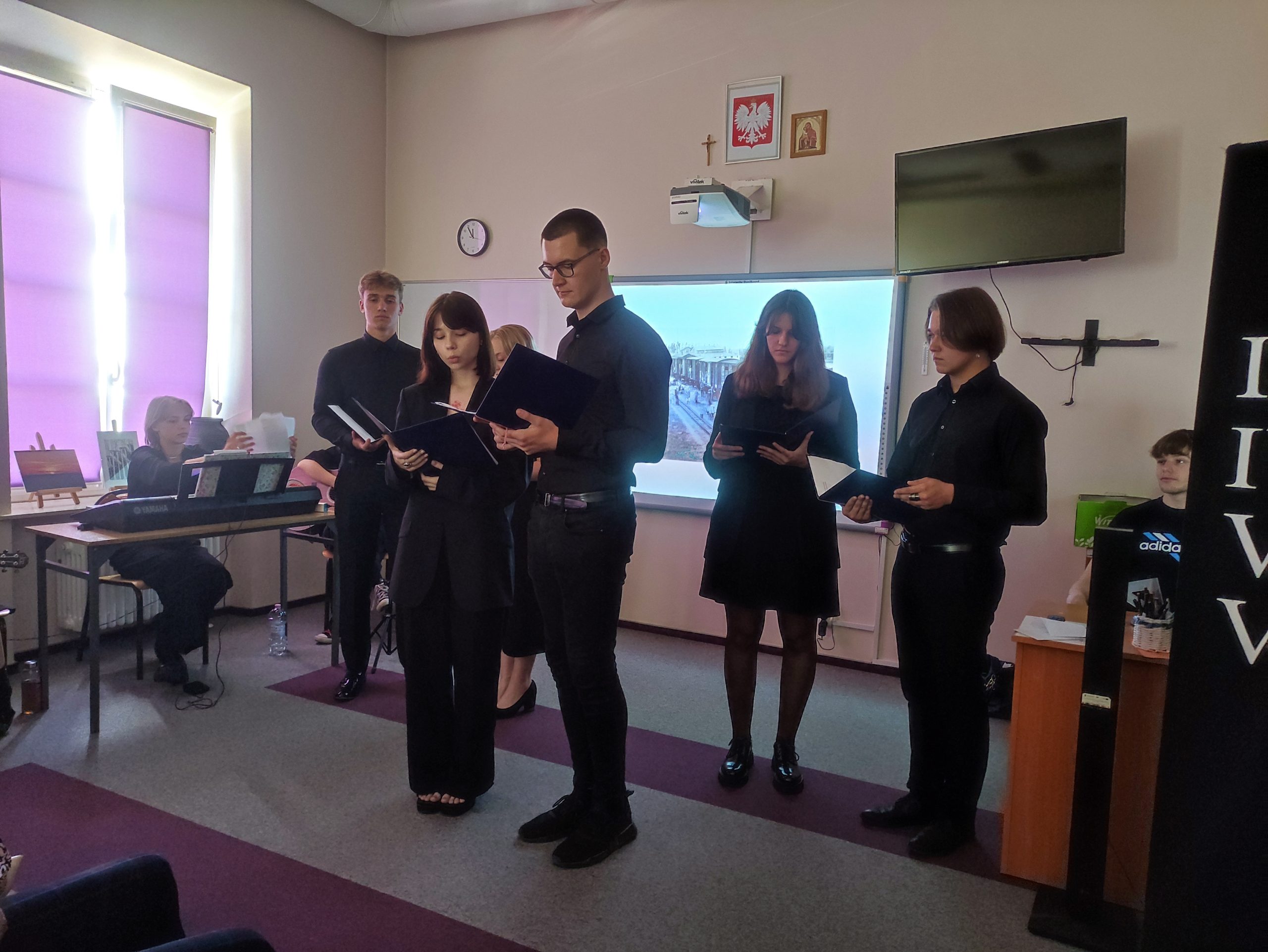 grupa uczniów ubranych na czarno trzyma w rękach podkładki, czyta