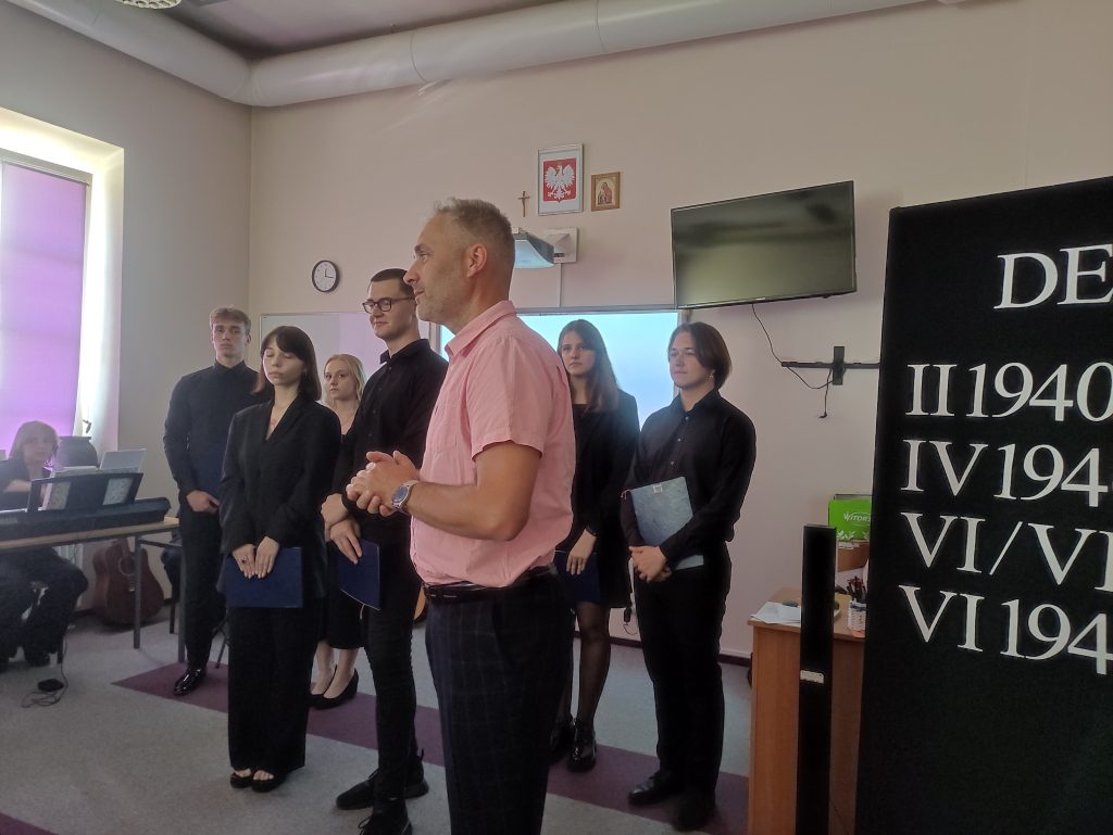 mężczyzna stoi bokiem, przemawia, w tle uczniowie